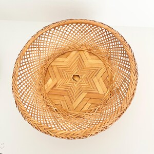 May include: A round, light brown woven basket featuring a complex lattice pattern on the sides and a geometric star-shaped design at the base. The basket is constructed from thin, flexible strips of natural material, creating an open weave.