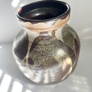 May include: A round ceramic vase with a wide mouth and a thick, flared rim. The exterior features a mottled glaze with overlapping shades of dark brown, cream, and muted olive green, creating a drip-like pattern that flows down the curved body.