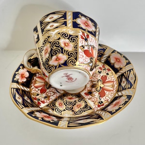 May include: A porcelain teacup and saucer set featuring a deep navy blue background with intricate gold filigree, white scrollwork, and orange floral motifs. The base of the cup displays a red maker's mark with the text Made in England.