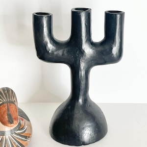 May include: A black, three-armed candelabra with a smooth, glossy finish and a wide, rounded base. The structure features a central pillar that branches into three distinct candle holders at the top, with visible hand-crafted texture.