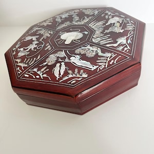 May include: An octagonal dark reddish-brown wooden box featuring intricate mother-of-pearl inlay on the lid. The lid displays detailed scenes of figures, trees, and a central circular motif, all crafted with iridescent shell accents.