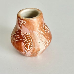 May include: A small, handcrafted ceramic vase with a bulbous shape and a wide opening. The vase is painted in shades of brown and peach, with white etched designs of geometric shapes and a stylized insect. The vase is approximately 4 inches tall.