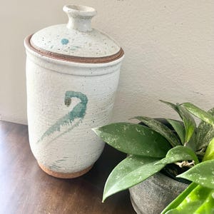 May include: A cylindrical ceramic canister with a speckled off-white glaze and a matching lid. The vessel features a hand-painted teal abstract design on the front and a small teal floral motif on the lid, with an exposed terracotta-colored base.