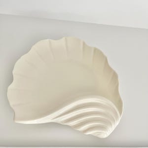 May include: A cream-colored ceramic dish shaped like a large seashell. The surface features a smooth, glossy finish with raised, curved ridges that radiate from one corner, mimicking the natural texture and form of a scallop shell.