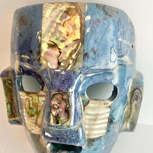 May include: A blue and iridescent mask with a face-like design. The mask features a mosaic pattern with blue, gold, and iridescent shell-like accents. The mask has eye and mouth openings, and is likely a decorative item.
