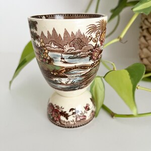 May include: A cream-colored ceramic egg cup featuring a detailed brown transferware landscape scene. The design depicts a river with a bridge, sailboats, mountains, and swans. The base is decorated with floral patterns in brown and muted red tones.