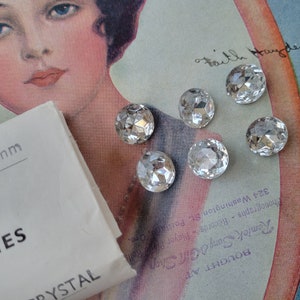 May include: A close-up of a package of 16 mm clear glass stones. The package is white with black text that reads "Size 16 mm" and "TONES CRYSTAL". The stones are scattered on a vintage magazine page with a woman's portrait.