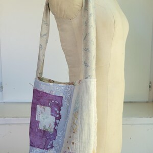 May include: A white fabric tote bag with a patchwork design featuring purple, blue, and white floral patterns. The bag has a long strap and is made from repurposed materials.