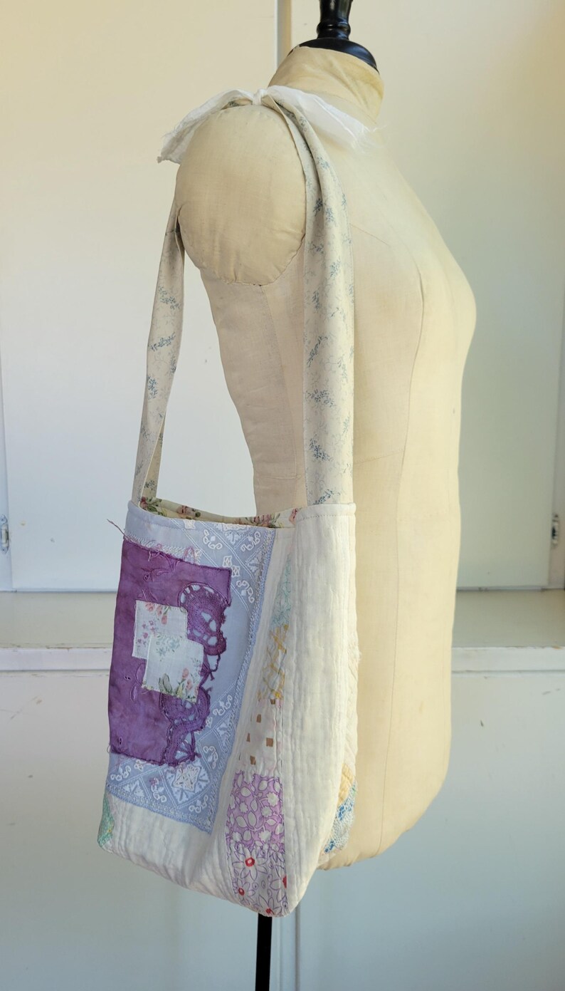 May include: A white fabric tote bag with a patchwork design featuring purple, blue, and white floral patterns. The bag has a long strap and is made from repurposed materials.