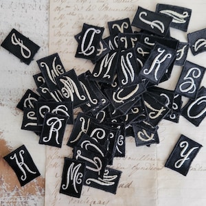 May include: A collection of black fabric patches embroidered with white cursive letters. The patches are rectangular with rounded corners. The letters include J, C, M, K, S, O, R, and Y. The patches are scattered on a vintage paper background.