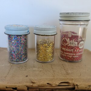 Puede incluir: Tres frascos de vidrio vintage llenos de purpurina. Un frasco contiene purpurina roja, otro dorada y el tercero una mezcla de colores. Los frascos tienen tapas de metal y se exhiben sobre una superficie marr&oacute;n.