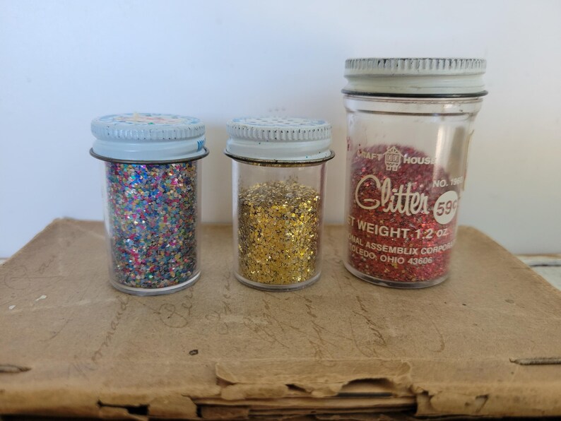 Puede incluir: Tres frascos de vidrio vintage llenos de purpurina. Un frasco contiene purpurina roja, otro dorada y el tercero una mezcla de colores. Los frascos tienen tapas de metal y se exhiben sobre una superficie marr&oacute;n.