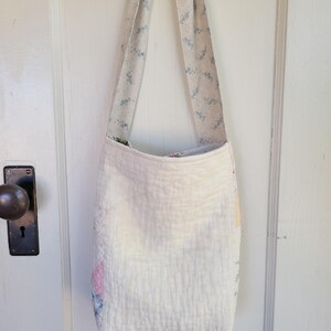 May include: A white quilted tote bag with a floral print on the straps. The bag is hanging on a wooden hanger.