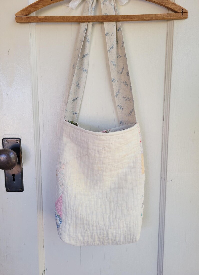 May include: A white quilted tote bag with a floral print on the straps. The bag is hanging on a wooden hanger.