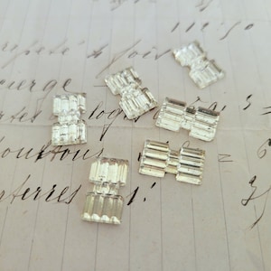 May include: A group of seven clear glass, decorative,  rectangular shaped,  faceted,  crystal-like,  jewelry findings.  The findings are scattered on a piece of lined paper with handwritten text.