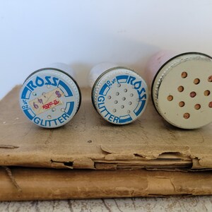 Puede incluir: Tres recipientes de purpurina vintage con tapas met&aacute;licas. Las tapas son blancas con texto azul que dice "ROSS GLITTER". Una tapa tiene una etiqueta de precio. Los recipientes est&aacute;n sobre una superficie de papel marr&oacute;n.