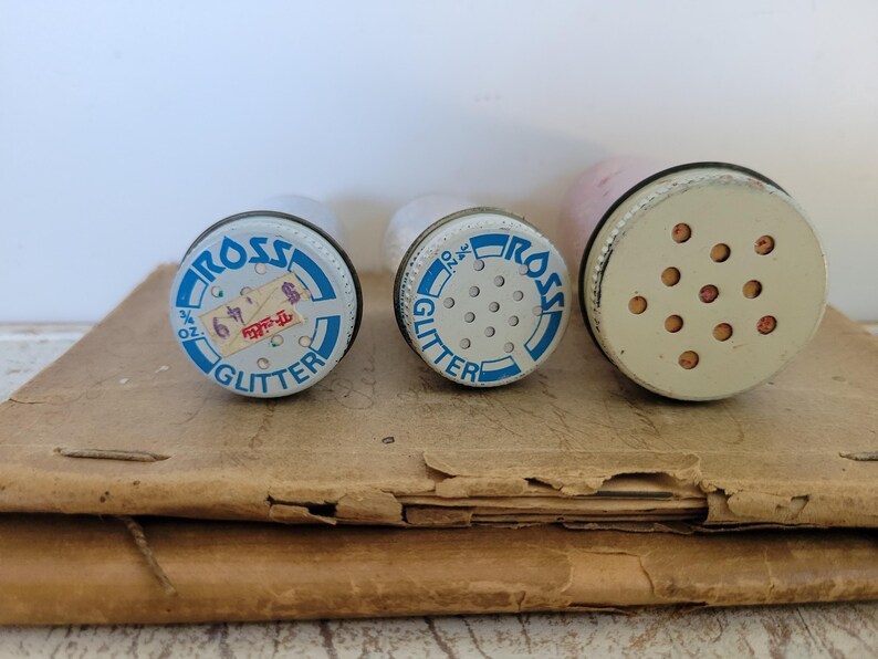 Puede incluir: Tres recipientes de purpurina vintage con tapas met&aacute;licas. Las tapas son blancas con texto azul que dice "ROSS GLITTER". Una tapa tiene una etiqueta de precio. Los recipientes est&aacute;n sobre una superficie de papel marr&oacute;n.