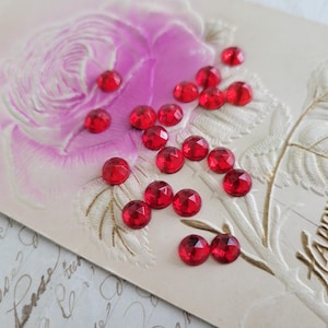 May include: Vintage postcard with a pink rose illustration and embossed gold accents. Scattered across the card are numerous faceted red gemstones. The words "Happy Returns" are printed in gold.