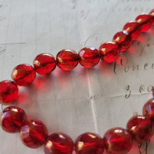 May include: A strand of faceted, round, red beads. The beads are strung together, creating a necklace or bracelet. The beads have a glossy finish and reflect light, with a vintage paper background.