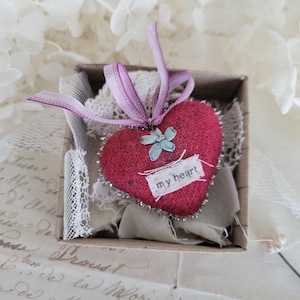 May include: A small, red heart-shaped ornament with the text "my heart" on a white tag, nestled in a cardboard box. The heart has a silver trim and a light blue flower accent. A purple ribbon is attached to the top.