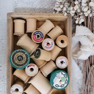 May include: A wooden box filled with vintage wooden spools of thread. Some spools have labels with text like "COATS & CLARK'S" and "BEST SIX CORD". The spools are various sizes and colors, with a natural wood finish. A bundle of white decorative stems is in the background.
