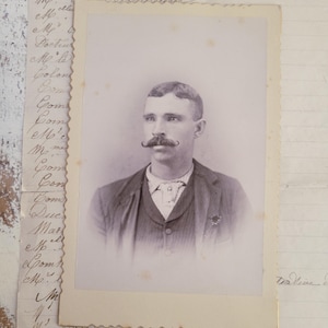 May include: A vintage sepia-toned portrait photograph of a person with a prominent handlebar mustache, wearing a collared shirt, vest, and jacket. The photograph is placed on top of a piece of aged, off-white paper featuring handwritten cursive text.