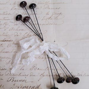 May include: A bundle of dark brown glass-headed pins tied with a white lace ribbon. The pins have long, thin black stems. The background is a vintage paper with faded handwriting.