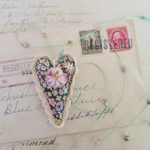 May include: A heart-shaped fabric pendant featuring a dark green background with a multi-colored floral pattern, edged with cream-colored crochet lace. A small pink fabric flower is attached to the center. It hangs from a thin light green cord.