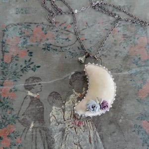 May include: A cream-colored crescent moon pendant necklace with a silver chain. The moon is adorned with small blue and purple fabric roses and a beaded edge. The background features a vintage floral and figure print.