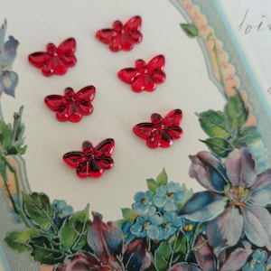 May include: Six red plastic butterfly-shaped buttons on a white background with a floral border.