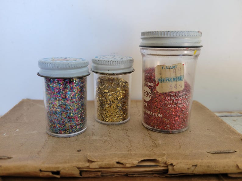 Puede incluir: Tres frascos de vidrio vintage llenos de purpurina. Los frascos tienen tapas de metal y est&aacute;n llenos de purpurina colorida, dorada y roja. Un frasco tiene una etiqueta de precio que dice "T-MART" y "59c".