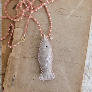 May include: A delicate necklace with a pink crocheted chain and a gray fabric fish pendant with white accents.