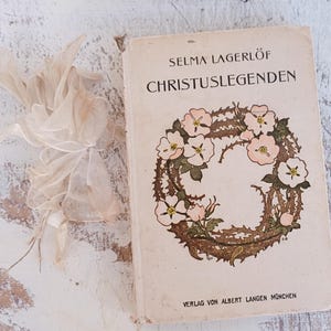 Puede incluir: Un libro vintage titulado "Selma Lagerlöf Christuslegenden" con una ilustración floral de una corona de rosas rosas en la portada. El libro está publicado por Verlag von Albert Langen München.