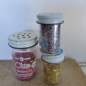 Puede incluir: Tres botes de purpurina vintage con tapas met&aacute;licas. Un bote contiene purpurina roja, otro purpurina de colores en forma de estrella y el tercero purpurina dorada. El bote con purpurina roja tiene el texto "Glitter" y "Craft House".