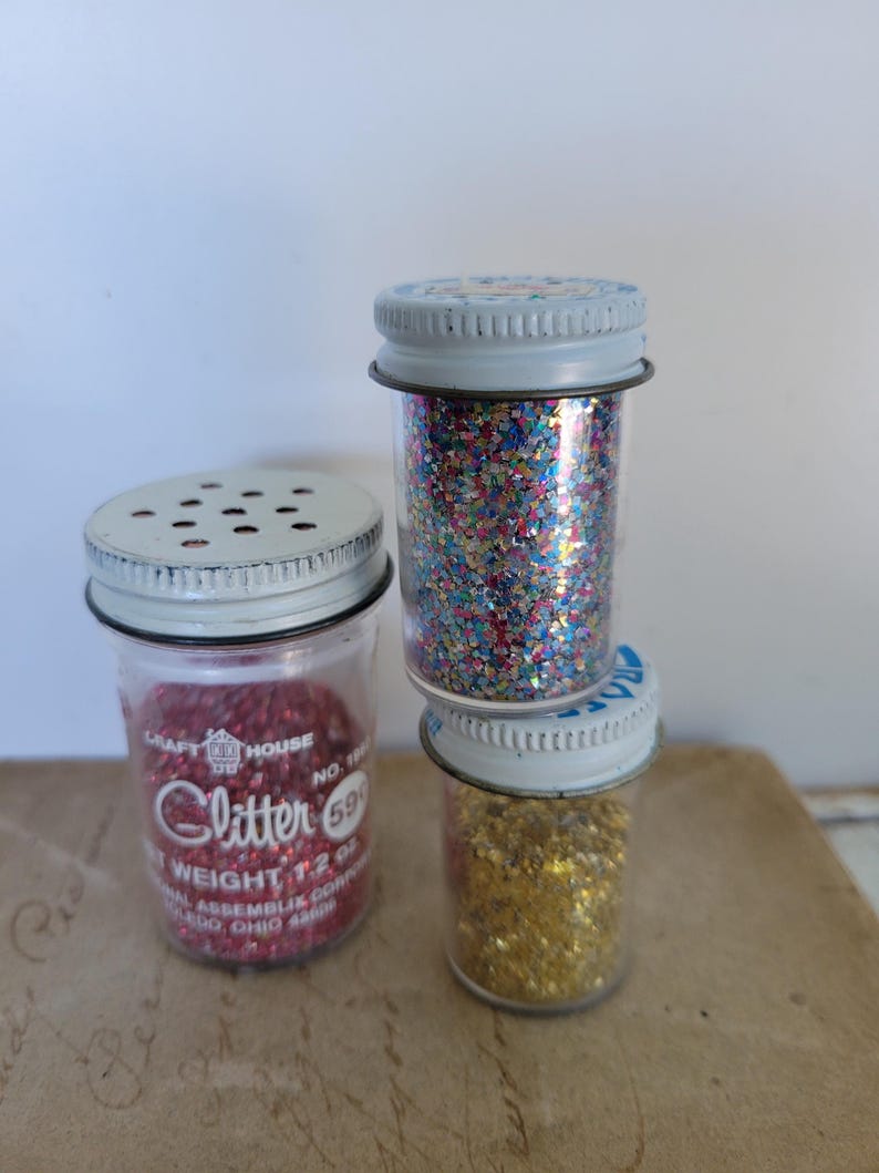 Puede incluir: Tres botes de purpurina vintage con tapas met&aacute;licas. Un bote contiene purpurina roja, otro purpurina de colores en forma de estrella y el tercero purpurina dorada. El bote con purpurina roja tiene el texto "Glitter" y "Craft House".