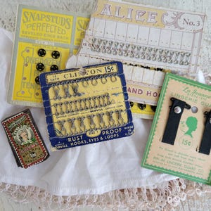 May include: A collection of vintage sewing notions, including cards of hooks, eyes, snaps, and lingerie guards. The items are displayed on a white cloth with lace trim, showcasing a variety of colors and packaging designs.
