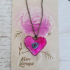 May include: A handmade necklace featuring a vibrant pink heart pendant with a small blue flower accent. The heart is outlined with a dark trim and hangs from a dark chain. The pendant rests on a vintage postcard with the words "HAPPY RETURNS".