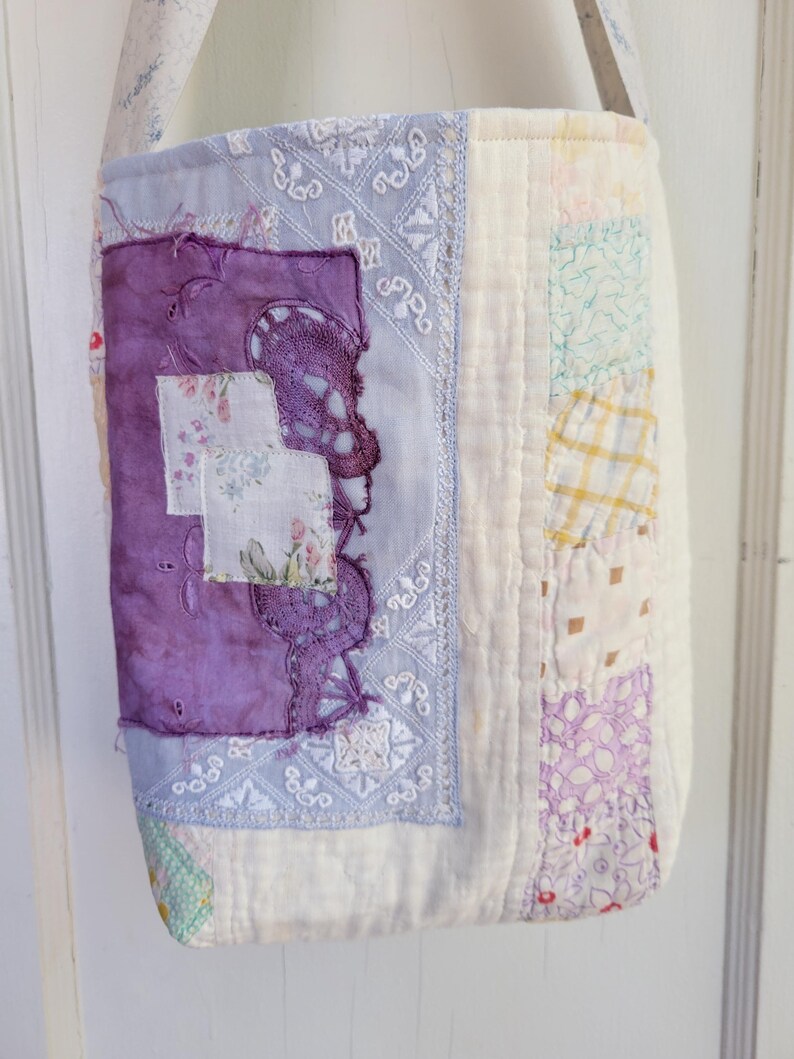 May include: A white patchwork tote bag with a purple and white floral design. The bag has a long strap and is made of various fabrics.