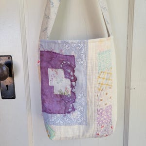 May include: A white patchwork tote bag with a purple and white floral design. The bag has a long strap and is made of fabric scraps.