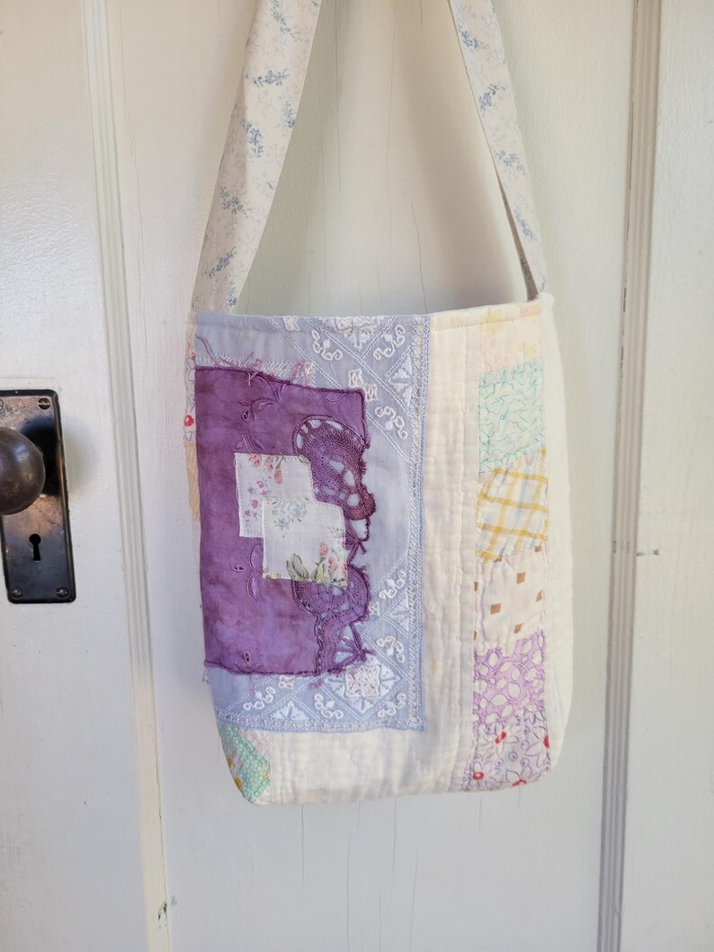 May include: A white patchwork tote bag with a purple and white floral design. The bag has a long strap and is made of fabric scraps.