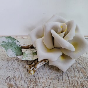 May include: A white porcelain rose with yellow accents and green leaves. The rose is lying on a white wooden surface.
