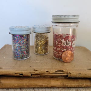 Puede incluir: Tres frascos de vidrio llenos de purpurina. Los frascos tienen tapas de metal y est&aacute;n llenos de purpurina de colores. Un frasco tiene purpurina roja y la etiqueta dice "Glitter". Una moneda est&aacute; al lado del frasco.