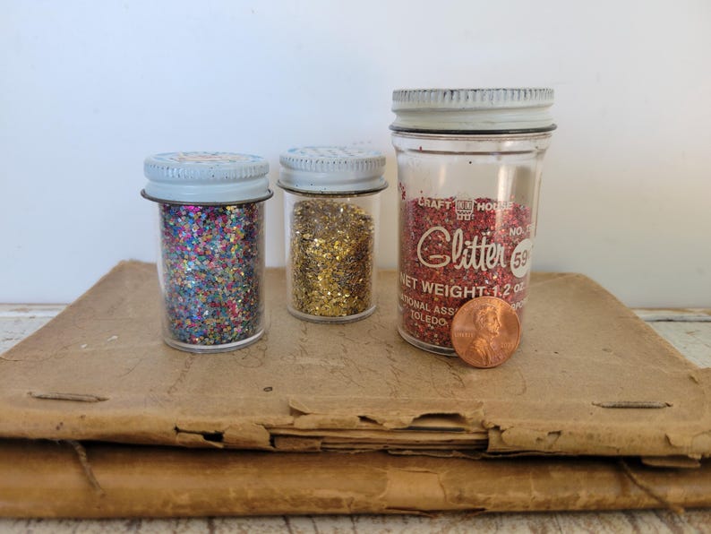 Puede incluir: Tres frascos de vidrio llenos de purpurina. Los frascos tienen tapas de metal y est&aacute;n llenos de purpurina de colores. Un frasco tiene purpurina roja y la etiqueta dice "Glitter". Una moneda est&aacute; al lado del frasco.