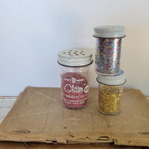 Puede incluir: Tres peque&ntilde;os frascos de vidrio llenos de purpurina roja, dorada y multicolor. Los frascos tienen tapas met&aacute;licas con peque&ntilde;os agujeros. El frasco de purpurina roja tiene el texto "Glitter 59" y "Peso neto 34g."