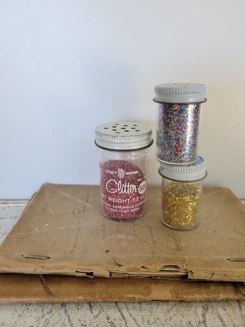 Puede incluir: Tres peque&ntilde;os frascos de vidrio llenos de purpurina roja, dorada y multicolor. Los frascos tienen tapas met&aacute;licas con peque&ntilde;os agujeros. El frasco de purpurina roja tiene el texto "Glitter 59" y "Peso neto 34g."