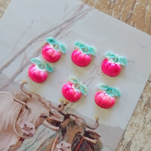May include: Six small, bright pink apple-shaped embellishments with green leaves are arranged on a vintage postcard featuring illustrations of two cows. The apples have a glossy finish and are likely decorative craft supplies.