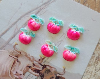 May include: Six small, bright pink apple-shaped embellishments with green leaves are arranged on a vintage postcard featuring illustrations of two cows. The apples have a glossy finish and are likely decorative craft supplies.