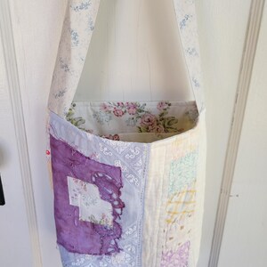 May include: A patchwork tote bag with a floral print lining. The bag is made from a variety of fabrics in shades of purple, blue, pink, and yellow. The strap is a light cream color with a blue floral pattern.