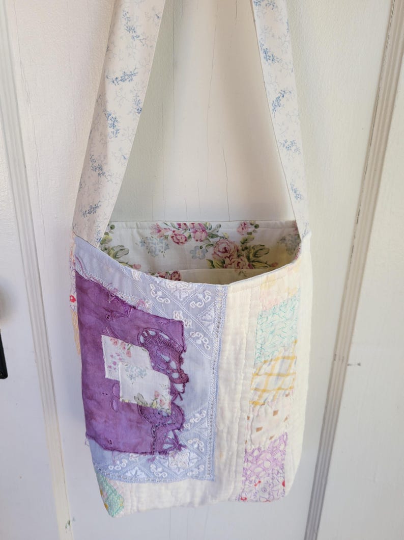 May include: A patchwork tote bag with a floral print lining. The bag is made from a variety of fabrics in shades of purple, blue, pink, and yellow. The strap is a light cream color with a blue floral pattern.