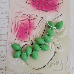 May include: A strand of ten small, smooth, light green beads shaped like elongated ovals or leaves, strung together on a thin white thread. The beads have a glossy finish and are arranged in a curved, organic pattern against a vintage postcard.